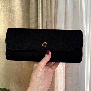 May include: A black velvet clutch with a gold heart-shaped clasp. The rectangular bag has a flap closure and a soft, textured appearance. The clutch is held up by a person's hand.
