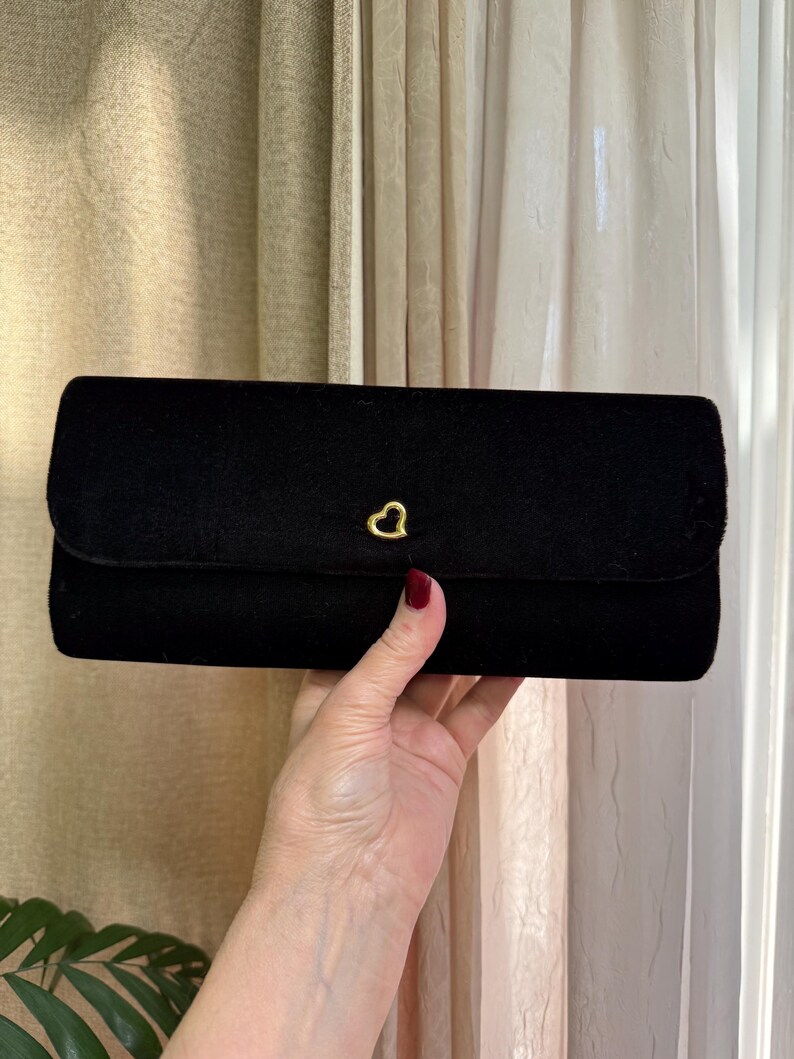 May include: A black velvet clutch with a gold heart-shaped clasp. The rectangular bag has a flap closure and a soft, textured appearance. The clutch is held up by a person's hand.