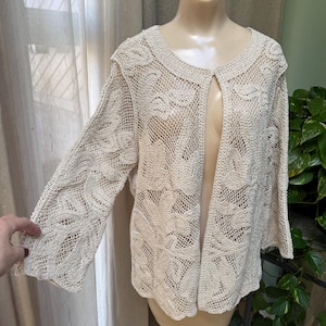 May include: A cream-colored crocheted cardigan with a floral pattern. The cardigan has a front button closure and long sleeves.