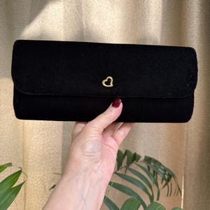May include: A black velvet clutch with a gold heart-shaped clasp. The rectangular bag has a flap closure and is held by a hand. The background is a neutral color.