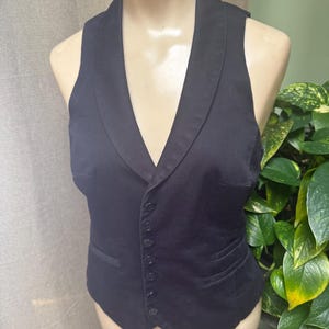 May include: A black vest with a notched lapel collar and a button-down front. The vest has a tailored fit and two front pockets. The garment is displayed on a mannequin.