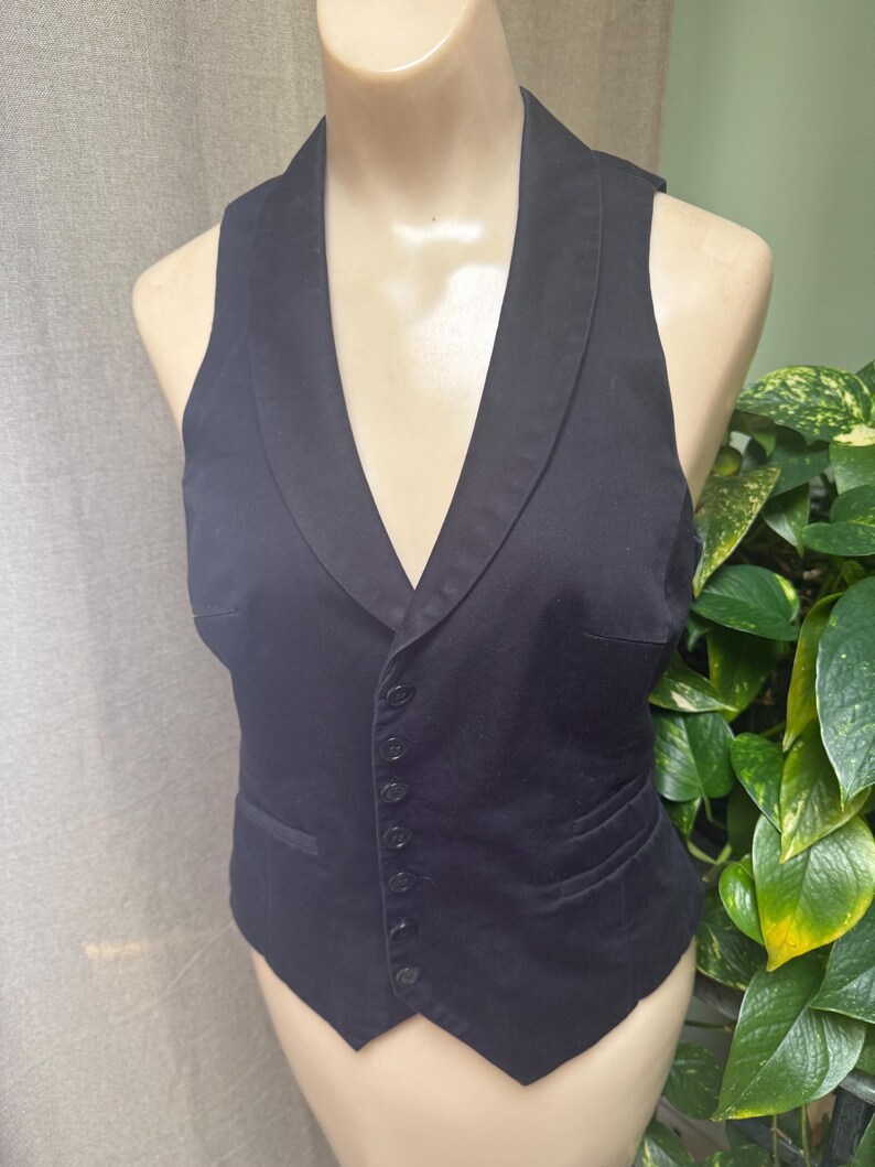 May include: A black vest with a notched lapel collar and a button-down front. The vest has a tailored fit and two front pockets. The garment is displayed on a mannequin.