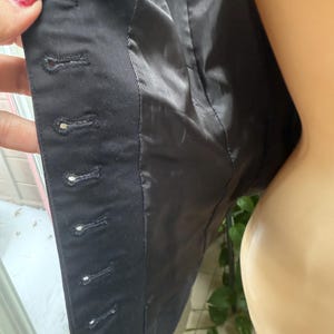 May include: Close-up of a black blazer's interior, showcasing the lining and button details. The blazer's fabric is dark, with visible stitching and buttonholes. The lining is a smooth, shiny black material.