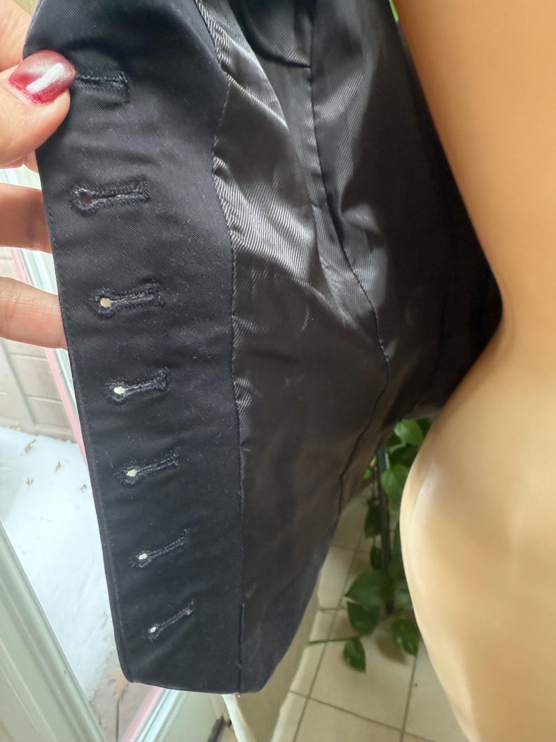 May include: Close-up of a black blazer's interior, showcasing the lining and button details. The blazer's fabric is dark, with visible stitching and buttonholes. The lining is a smooth, shiny black material.