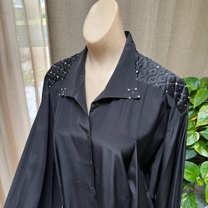 May include: Black button-down shirt with long sleeves. The shoulders feature quilted detailing and are embellished with small, sparkling rhinestones. The shirt has a classic collar and a button-up front.