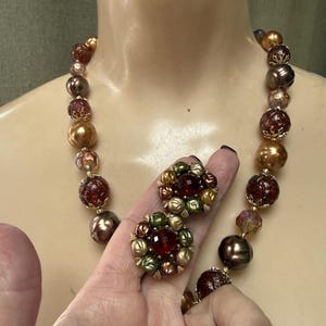 May include: A vintage necklace and matching clip-on earrings set. The necklace features a strand of various sized beads in shades of gold, brown, and burgundy. The earrings are round with a central red stone and surrounded by green and gold accents.