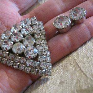 Pu&ograve; includere: Un fermaglio d'argento con un grande gruppo di strass trasparenti a forma triangolare. Sono raffigurati anche due piccoli orecchini di strass trasparenti.