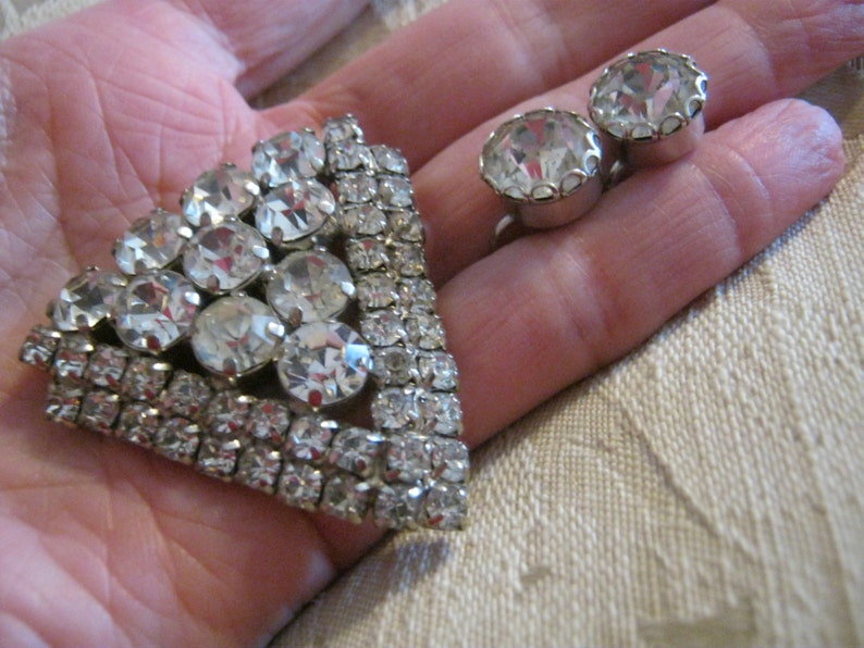 Pu&ograve; includere: Un fermaglio d'argento con un grande gruppo di strass trasparenti a forma triangolare. Sono raffigurati anche due piccoli orecchini di strass trasparenti.