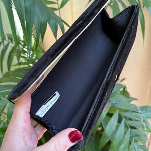 May include: Black velvet clutch with an open flap, revealing a black interior and a tag that reads "VICTORIA'S SECRET". The clutch is held open, showcasing its interior. The background features green foliage.