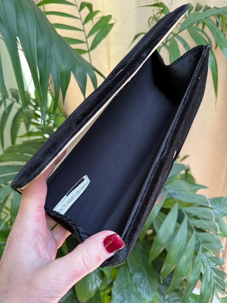 May include: Black velvet clutch with an open flap, revealing a black interior and a tag that reads "VICTORIA'S SECRET". The clutch is held open, showcasing its interior. The background features green foliage.