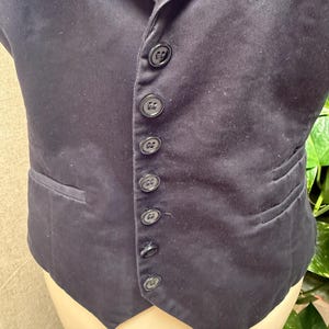 May include: A dark navy vest with a notched lapel and a row of six black buttons down the front. The vest has a single pocket on the left side and a tailored fit. The fabric appears to be a smooth, woven material.