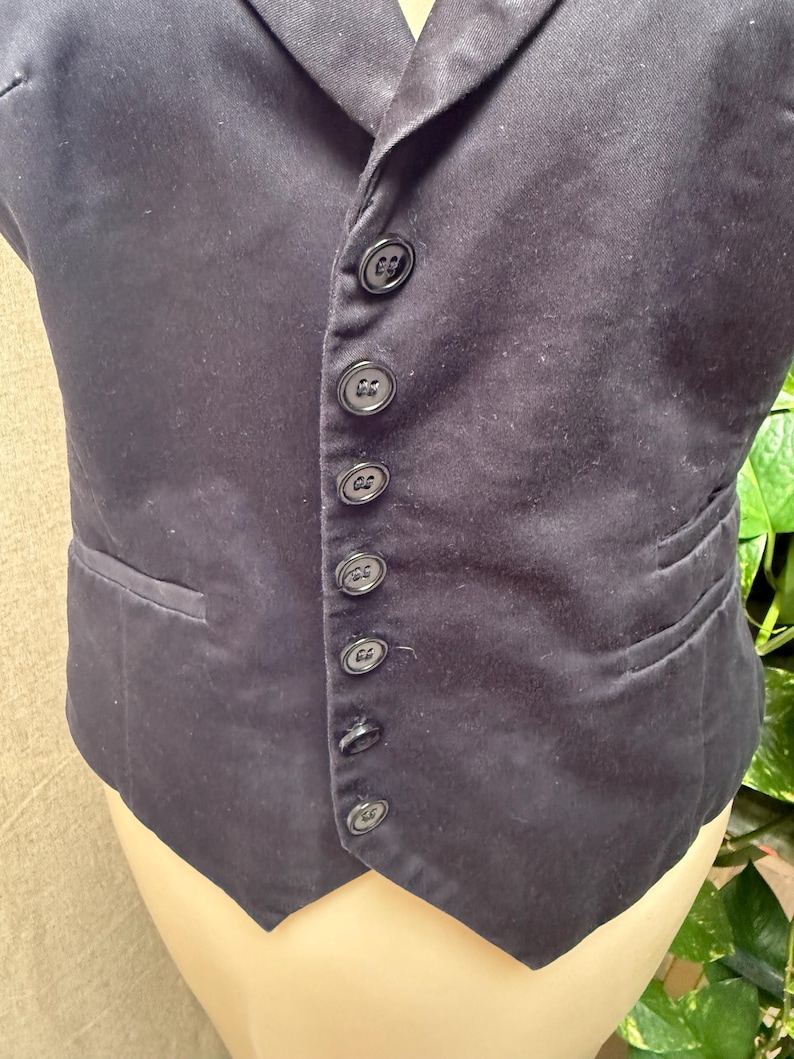May include: A dark navy vest with a notched lapel and a row of six black buttons down the front. The vest has a single pocket on the left side and a tailored fit. The fabric appears to be a smooth, woven material.