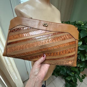 May include: A tan leather clutch with a unique design. The center features a patchwork of textured, patterned strips in shades of brown and tan. The top and bottom are smooth tan leather, with a gold-tone clasp.