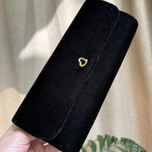 May include: A black velvet clutch with a gold heart-shaped clasp. The rectangular clutch has a rounded flap and a soft, textured appearance. The clasp is centered on the front of the clutch.