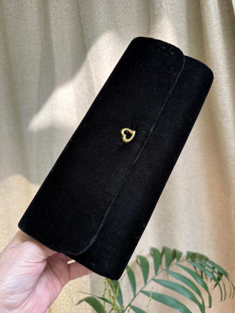 May include: A black velvet clutch with a gold heart-shaped clasp. The rectangular clutch has a rounded flap and a soft, textured appearance. The clasp is centered on the front of the clutch.