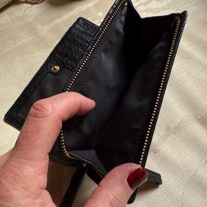May include: Black leather wallet with a gold zipper and a small pocket on the outside. The wallet is open and the inside is lined with black fabric.
