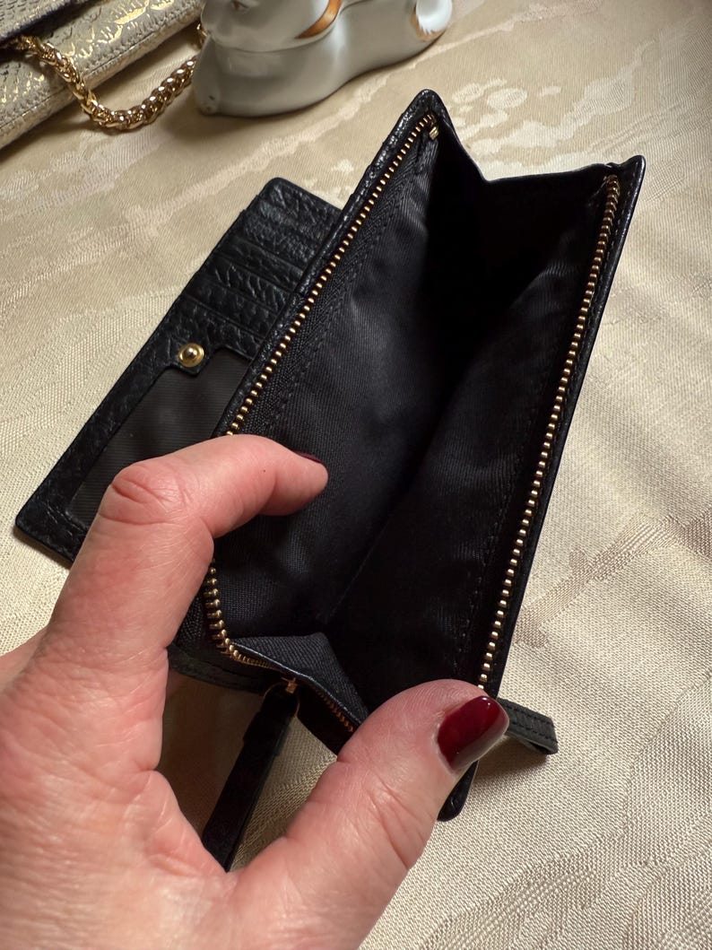 May include: Black leather wallet with a gold zipper and a small pocket on the outside. The wallet is open and the inside is lined with black fabric.