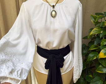 May include: A white long-sleeved blouse featuring intricate white embroidery on the cuffs, paired with a black fabric sash tied at the waist. A gold-toned necklace with a large oval pendant rests at the neckline of the garment.
