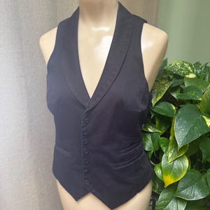 May include: A black vest with a notched lapel collar and a button-down front. The vest has a tailored fit and a classic design. The vest is displayed on a mannequin.