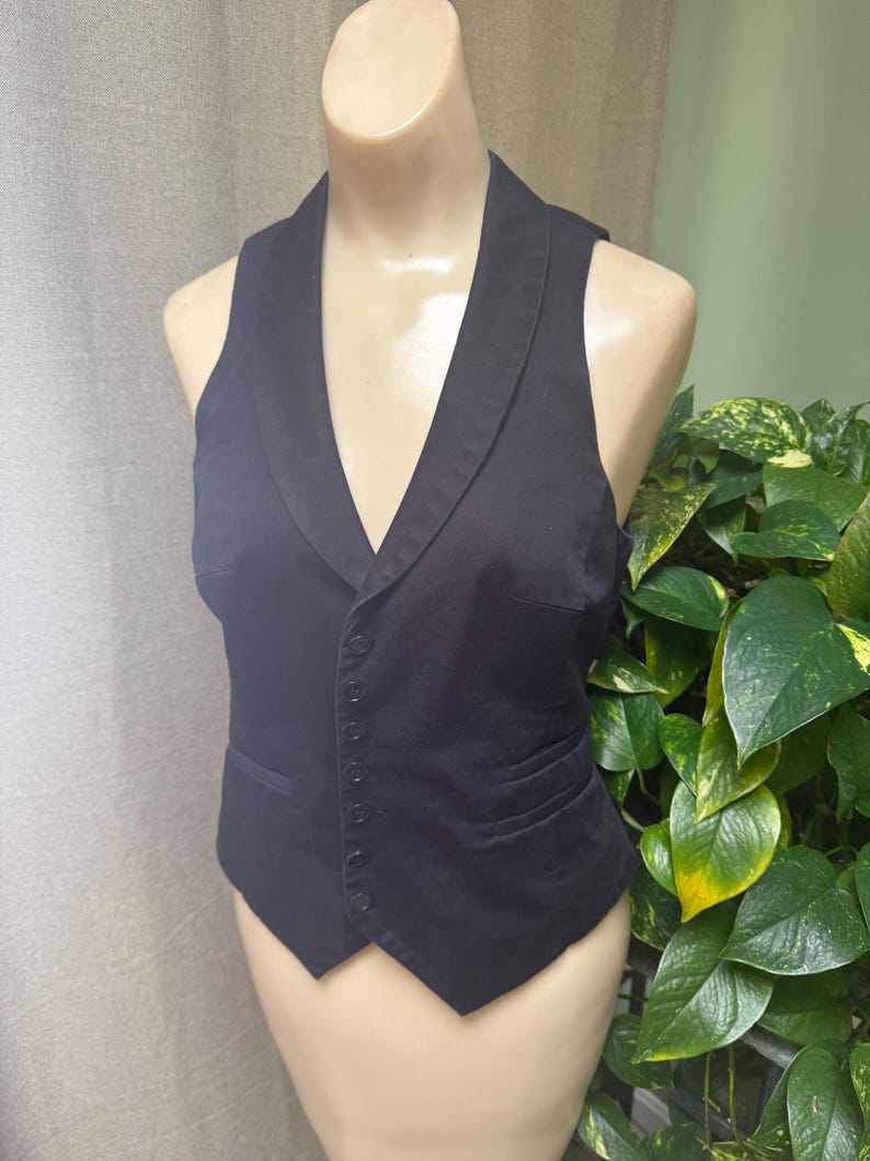 May include: A black vest with a notched lapel collar and a button-down front. The vest has a tailored fit and a classic design. The vest is displayed on a mannequin.