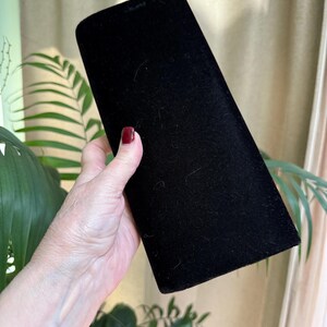 May include: A black velvet jewelry box with a rectangular shape and rounded top. The box is held in a person's hand, with a background of green plants and a beige curtain.