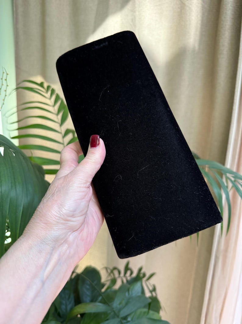 May include: A black velvet jewelry box with a rectangular shape and rounded top. The box is held in a person's hand, with a background of green plants and a beige curtain.