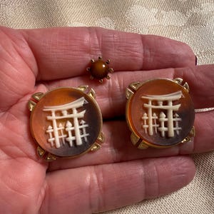 May include: A set of vintage jewelry, including a pair of round earrings and a small brooch. The earrings and brooch feature a gold-tone setting with a brown and white design. The earrings have a Japanese Torii gate design.