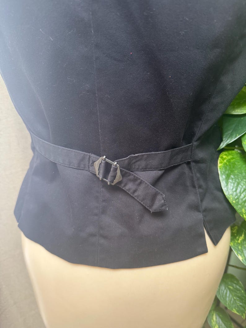 May include: Black vest with an adjustable belt and buckle. The vest is made of a smooth fabric and has a tailored fit. The buckle is silver-toned and adds a decorative element to the back of the vest.