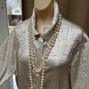 May include: A white satin blouse with a textured pattern and a long pearl necklace. The necklace is made of large, round pearls and is worn as a double strand.