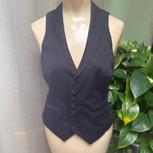 May include: A black vest with a shawl collar and a button-up front. The vest has a tailored fit and two front pockets. It is displayed on a mannequin. The background includes a neutral curtain and green plants.