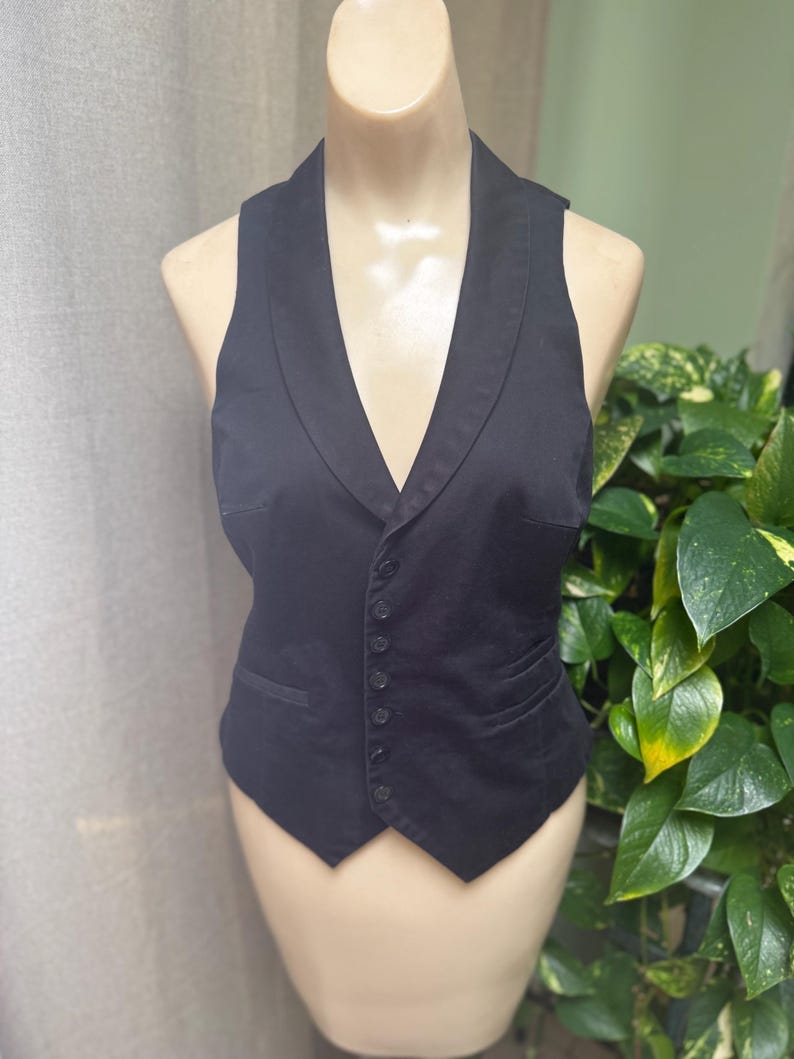 May include: A black vest with a shawl collar and a button-up front. The vest has a tailored fit and two front pockets. It is displayed on a mannequin. The background includes a neutral curtain and green plants.