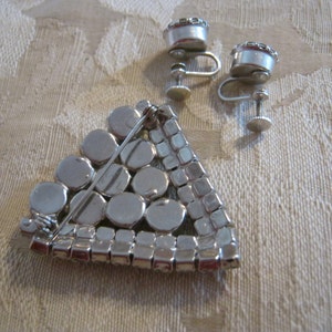 Pu&ograve; includere: Un broche triangolare color argento con un design geometrico di strass rotondi e quadrati. Il broche ha una chiusura a spilla ed &egrave; accompagnato da un paio di orecchini a clip abbinati.