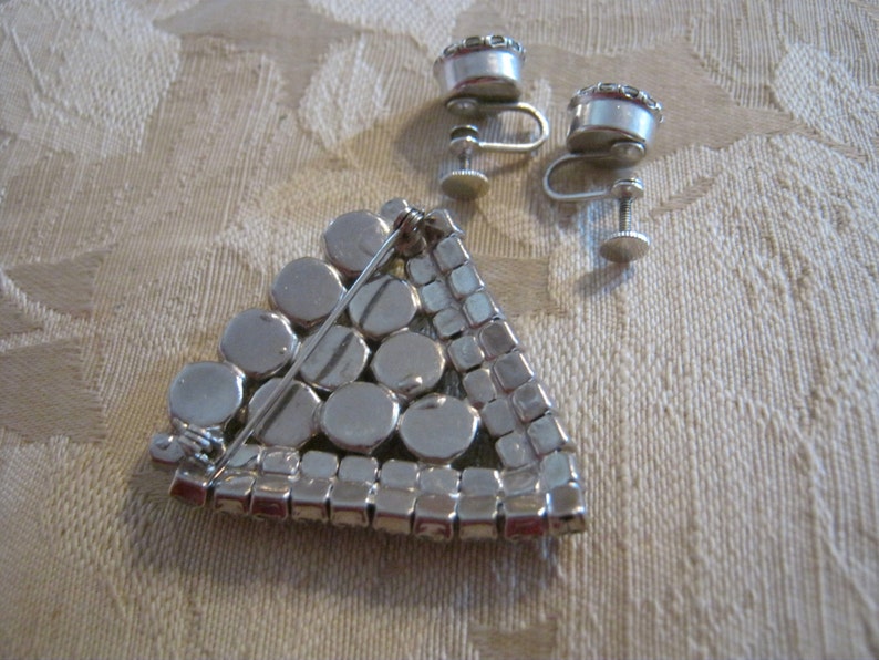 Pu&ograve; includere: Un broche triangolare color argento con un design geometrico di strass rotondi e quadrati. Il broche ha una chiusura a spilla ed &egrave; accompagnato da un paio di orecchini a clip abbinati.