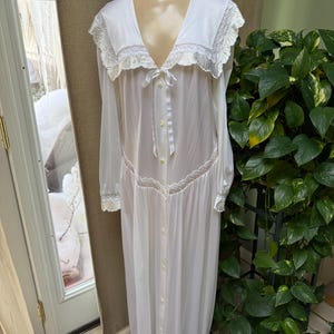May include: A long, white nightgown with a V-neckline and long sleeves. The nightgown features delicate lace trim around the neckline, cuffs, and waist. It has a button-down front and a ribbon tie at the bust. The garment is displayed on a mannequin.