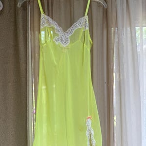 May include: A neon yellow chemise featuring a V-neckline with white lace trim, thin spaghetti straps, and a side slit at the hem accented with white lace and a small peach-colored bow. The garment is made of a sheer, lightweight fabric.