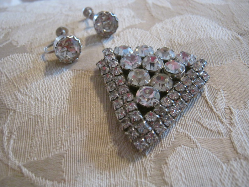 Pu&ograve; includere: Un set di gioielli vintage con una spilla triangolare con strass trasparenti e un paio di orecchini abbinati con pietre trasparenti.