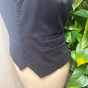 May include: A black vest with a notched lapel and a row of buttons down the front. The vest has two pockets and a tailored fit. The fabric appears to be a smooth, woven material. The vest is displayed on a mannequin.