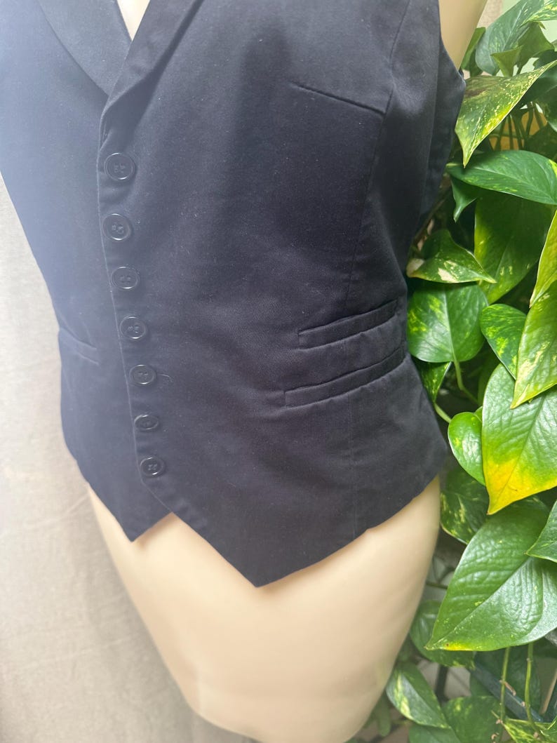 May include: A black vest with a notched lapel and a row of buttons down the front. The vest has two pockets and a tailored fit. The fabric appears to be a smooth, woven material. The vest is displayed on a mannequin.