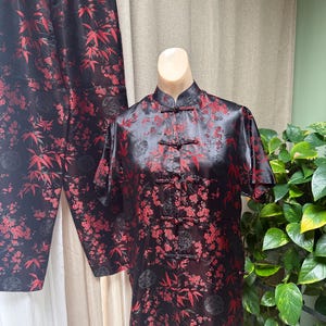 May include: A two-piece pajama set featuring a short-sleeved top and matching pants in black satin fabric. The garment is decorated with a repeating pattern of red bamboo leaves and floral blossoms, accented by traditional knotted fabric closures.