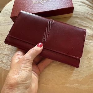 May include: A burgundy leather wallet with a textured flap and a decorative emblem. The wallet is open, revealing the interior. A matching burgundy box with the brand name "Buxton" is in the background.