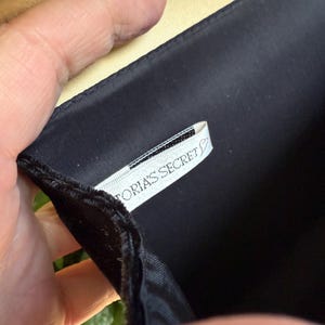 May include: Black satin clutch with a gold interior. The clutch is held open, revealing a white tag with the text "Victoria's Secret". The clutch has a simple, elegant design.