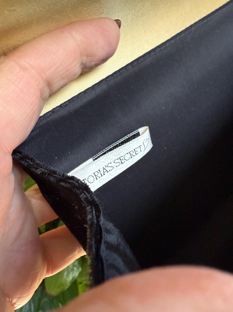 May include: Black satin clutch with a gold interior. The clutch is held open, revealing a white tag with the text "Victoria's Secret". The clutch has a simple, elegant design.