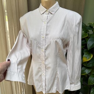 May include: A light pink long-sleeve button-down shirt with a pointed collar and a textured, crinkled fabric finish. The shirt features a central vertical button placket, structured shoulders, and cuffs with single buttons on each sleeve.