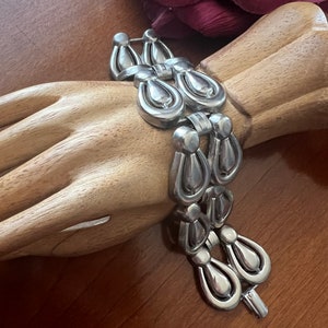 May include: A silver bracelet with a repeating pattern of teardrop-shaped links. The bracelet is on a wooden mannequin hand.