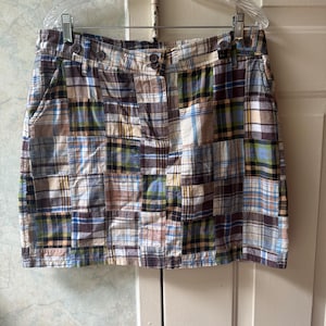 May include: A plaid patchwork skirt featuring a variety of square fabric panels in shades of brown, blue, green, and white. The skirt has a waistband with belt loops, a button closure, and side pockets, all displayed on a clear plastic hanger.