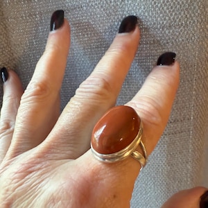 May include: A silver ring featuring a large, oval, reddish-brown gemstone. The ring has a split shank design and a polished finish. The gemstone is set in a bezel setting, and the ring is shown on a hand with dark nail polish.