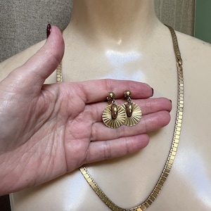 May include: A gold chain necklace and matching earrings. The necklace is made of small, square-shaped links. The earrings feature a round, fan-shaped design.