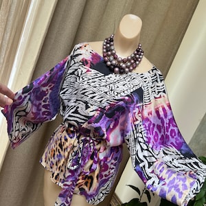 May include: A purple, black, and white patterned blouse with a tie belt. The blouse has a scoop neck and 3/4 sleeves. The blouse is on a mannequin wearing a multi-colored beaded necklace.