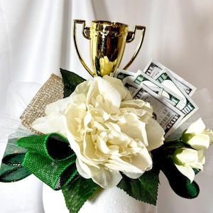 May include: A white flower arrangement with green leaves and a gold trophy on top. The arrangement is decorated with dollar bills.