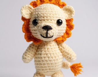 Puede incluir: Un juguete de león de ganchillo hecho a mano. El león tiene un cuerpo de color crema, una melena naranja y una pequeña cola naranja. Tiene ojos negros y una nariz y boca negras. El juguete está de pie.